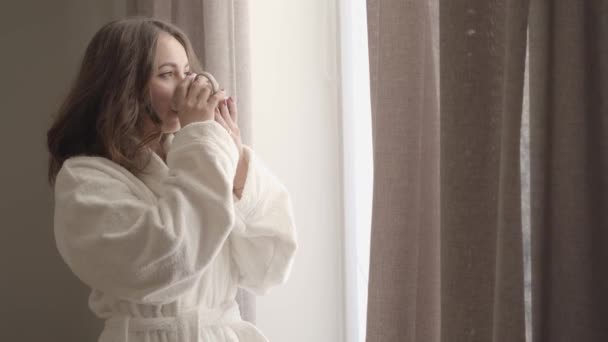 Porträtt av charmig ung dam i vit mjuk badrock njuter solig morgon hemma. Söt vit brunett kvinna tittar ut genom fönstret och dricker kaffe från koppen. — Stockvideo