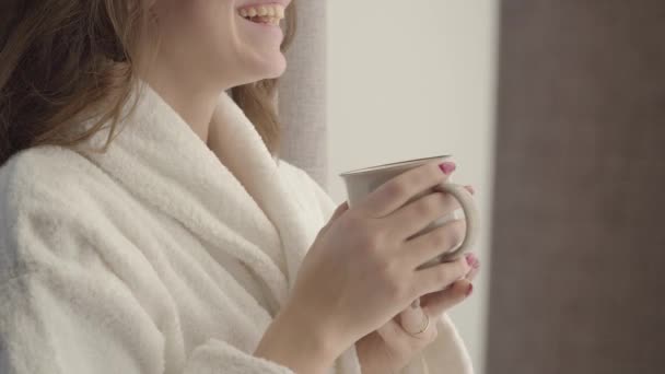 Primer plano de una joven caucásica irreconocible en albornoz blanco sosteniendo una taza de café y riendo. Joven morena positiva disfrutando de la mañana soleada en casa. Diversión, relajación, estilo de vida. ocio . — Vídeos de Stock