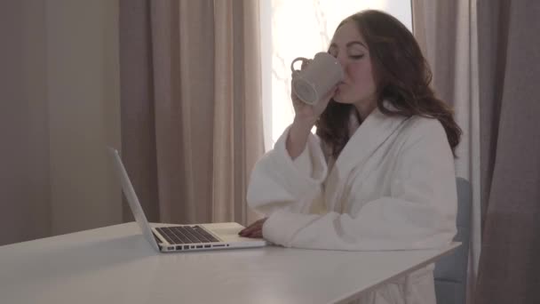 Mulher morena caucasiana atraente bebendo café e usando laptop pela manhã. Retrato de uma jovem encantadora sentada em casa, navegando na Internet e sorrindo. Mídia social, estilo de vida, online . — Vídeo de Stock