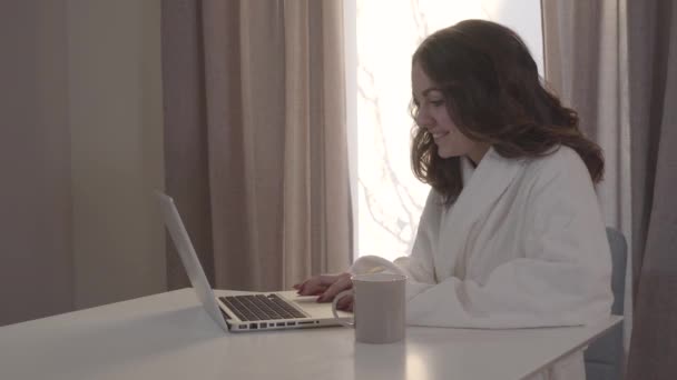 Mujer positiva caucásica mirando a la pantalla del ordenador portátil y sonriendo. Jovencita en albornoz blanco usando las redes sociales por la mañana. Chica bonita descansando en casa . — Vídeos de Stock