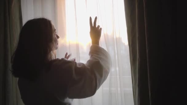 Retrato da jovem caucasiana olhando pela janela no início da manhã. Mulher bonita morena tocando luz cortina branca e sorrindo . — Vídeo de Stock