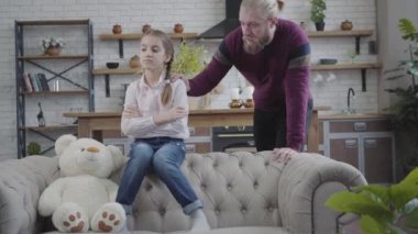 Yetişkin, yakışıklı, beyaz bir adam kızlarının omzunda elini tutuyor ve konuşuyor. Kanepede elleri çaprazlanmış oyuncak ayıyla oturan üzgün çocuk. Ergenlik, aile, bakım.