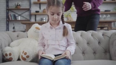 Sevimli beyaz kız portresi kanepeye oturmuş oyuncak ayıyla kitap okuyor ve elleriyle gözlerini kapatıyor. Neşeli adam sevimli kızıyla vakit geçiriyor. Mutluluk.