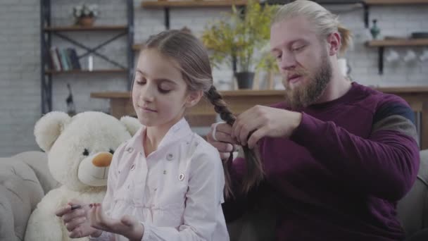 Молодой бородатый белый отец плетет косички дочерям, пока она подшивает гвозди и отворачивается. Взрослый мужчина и симпатичная девушка-подросток проводят свободное время вместе дома. Отцовство, забота, семья . — стоковое видео