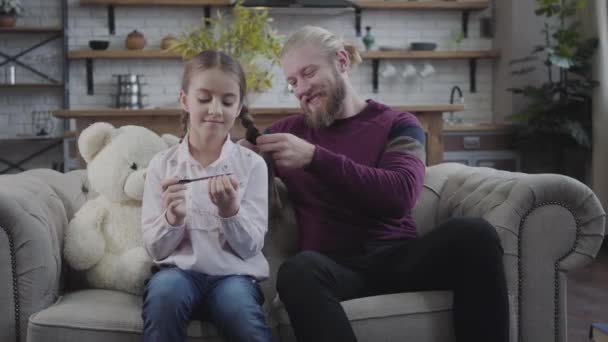 Portrait of handsome Caucasian man sitting on sofa with daughter and braiding pigtails. Happy girl spending time with father at home. Fatherhood, care, parenthood. — ストック動画