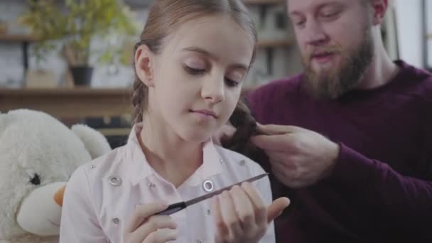 Cara de cerca de la niña caucásica reflexiva limando las uñas como padre sonriente trenzando sus coletas al fondo. Hermosa hija adolescente pasar tiempo con los padres en el interior. Cuidado, paternidad . — Vídeos de Stock