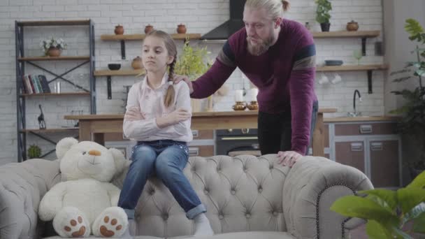 Adult handsome Caucasian man holding hand on daughters shoulder and talking. Sad child sitting on couch with teddy bear with hands crossed. Adolescence, family, care. — Stock Video