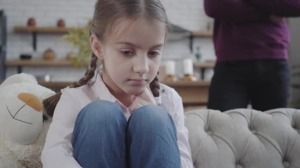 Close-up af søde kaukasiske brunette pige med flettet rottehaler sidder på sofaen og kramme knæ. Ugenkendelig far gestikulerer i baggrunden. Ungdom, adfærd, familieforhold . – Stock-video