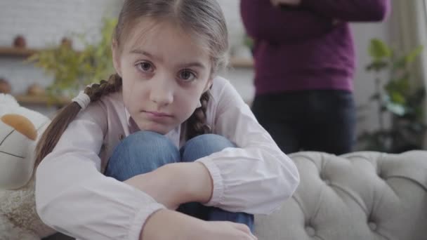 Close-up de menina branca bonita com olhos castanhos abraçando joelhos e olhando para a câmera. Homem irreconhecível ao fundo com as mãos cruzadas. Paternidade, adolescência . — Vídeo de Stock