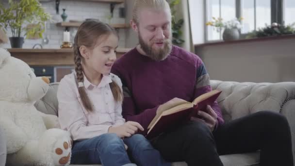 Närbild av glad vit man läser bok med tonårsdotter. Söt flicka med flätade flätor spenderar fritid med far hemma. Faderskap, omsorg, hobby, tonåren. — Stockvideo