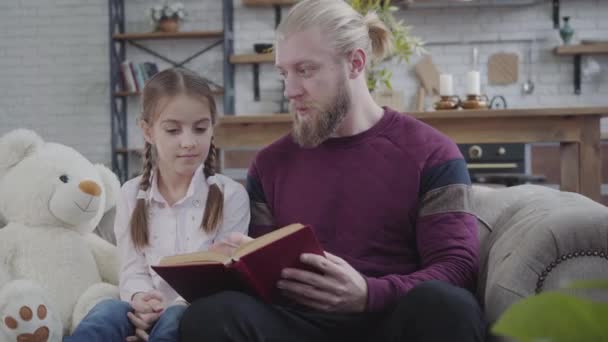 Fiatal kaukázusi férfi ül könyvvel és tinédzser lánnyal beszél. Barna lány rázza a fejét, igen. Család, vicces, szabadidő, életmód. — Stock videók