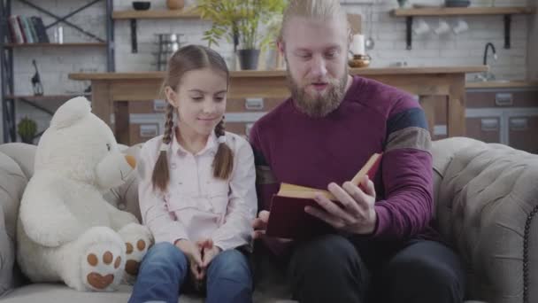 Père caucasien positif tournant la page et touchant le nez des filles. Adolescente mignonne souriante et regardant l'homme. Un adulte éduquant son enfant à la maison. Fun, passe temps, style de vie . — Video