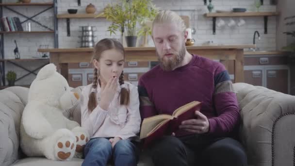 Retrato de una chica caucásica sorprendida escuchando a su padre leyendo su libro. Hombre positivo tocando la nariz de las hijas con la mano. Familia divirtiéndose en casa. paternidad, pasatiempo, estilo de vida . — Vídeos de Stock