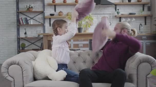 Joyful Caucasian father and daughter sitting on couch and fighting with pillows. Cheerful man and teenage girl having fun at home on weekends. Leisure, happiness, enjoyment. — ストック動画