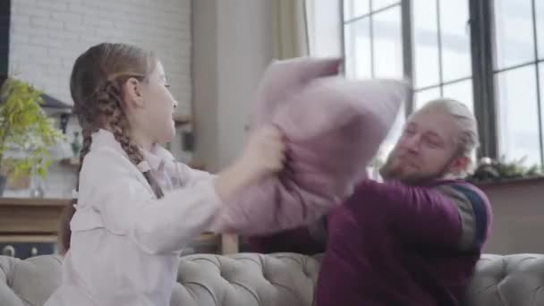 Close-up of laughing Caucasian teenage girl fighting pillows with father. Young man having fun with daughter on weekends. Camera moving around people slowly from left to right. — Stock Video