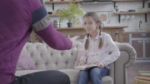 Retrato de linda chica caucásica guardando el libro, tomando la mano de los padres y de pie desde el sofá. Un hombre irreconocible bailando con su hija en casa. Felicidad, ocio, paternidad . — Vídeos de Stock