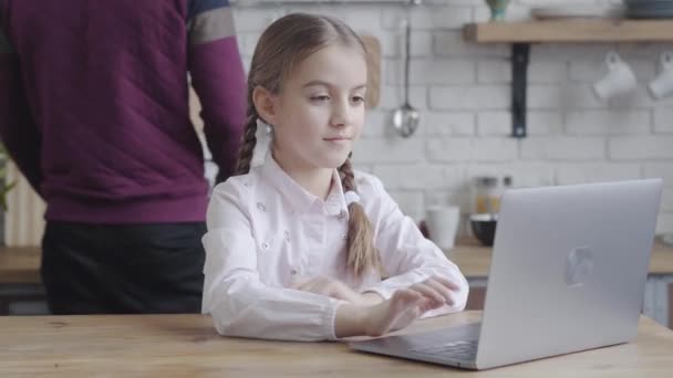 Primer plano de hermosa chica adolescente caucásica escribiendo en el teclado del ordenador portátil, mientras que el hombre irreconocible cocinar en el fondo. Hija navegando por Internet y esperando el desayuno. Adolescencia, mañana . — Vídeo de stock