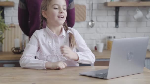 Portrait of cute Caucasian female teenager looking at laptop screen, holding head with hand and smiling. Pretty girl resting at kitchen while father cooking at the background. Morning, breakfast — ストック動画