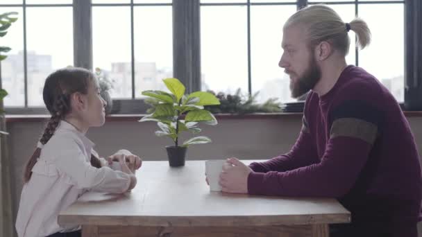 Jonge blanke man en tienermeisje zitten aan tafel en praten. Vader en dochter brengen de ochtend samen door thuis met een kop thee of koffie. Vriendelijke familie communiceert binnen. — Stockvideo