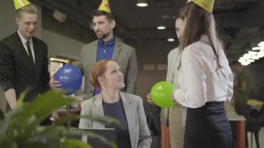 Parti şapkalı beyaz iş arkadaşları kadın iş arkadaşlarının etrafında balonlarla dikilip konuşuyorlar. Mutlu orta yaşlı kadın iş yerinde doğum gününü kutluyor. Olay, parti, mutluluk, kutlama.
