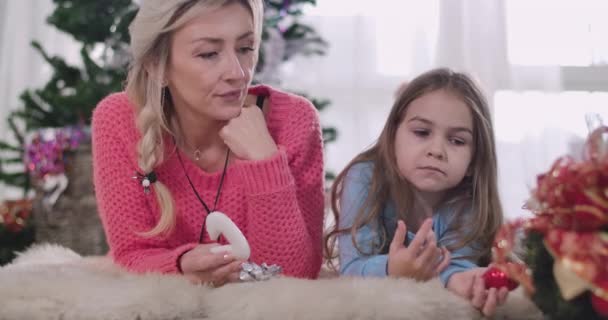 Retrato de una joven madre e hija caucásicas acostadas sobre una alfombra suave y comiendo galletas. Mujer adulta y niña descansando en el fondo del árbol de Navidad. Sede del cine 4k ProRes . — Vídeo de stock