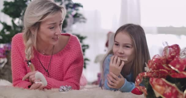 Positiv vit mor och dotter skrattar högt som att äta kakor hemma. Kvinna och flicka ligger på mjuk vit matta i bakgrunden av julgranen. Cinema 4k Prores Hq. — Stockvideo