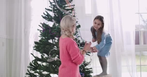 Retrato de jovem caucasiana ajudando menina bonita morena para decorar a árvore de Natal. Mãe e filha se preparando para a véspera de Ano Novo juntos em casa. Cinema 4k ProRes HQ . — Vídeo de Stock