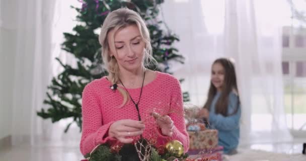 Retrato de la hermosa mujer rubia caucásica eligiendo decoraciones de Año Nuevo, chica morena en el fondo mirando a mamá, de pie, y guirnaldas colgantes en el árbol de Navidad. Sede del cine 4k ProRes . — Vídeo de stock