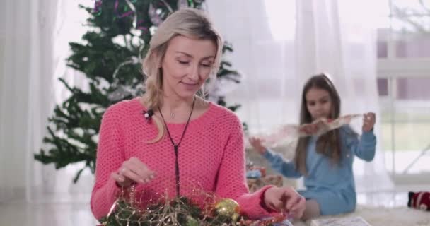 Brunette fille caucasienne choisissant décorations de Noël à l'arrière-plan comme sa mère assise au premier plan et souriant. Femme aidant sa fille à se préparer pour le réveillon du Nouvel An. Siège social Cinema 4k ProRes . — Video