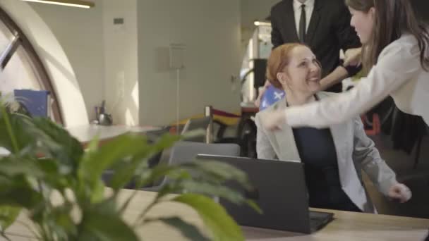 Jeune femme blanche attrayante assise à la table avec un ordinateur portable que ses collègues viennent avec des ballons portant des chapeaux de fête. Femme rousse de bureau célébrant son anniversaire à Worplace. Espace ouvert . — Video