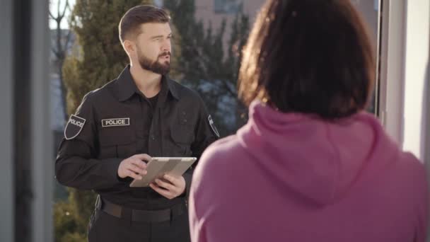 Jeune policier caucasien debout à la porte d'entrée et posant des questions. Femme brune montrant loin avec la main. Inspecteur enquêtant sur le crime. Témoignage oculaire, protection, interrogatoire . — Video