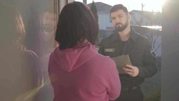 Egy fiatal kaukázusi rendőr portréja, aki az üvegajtó mögött áll táblagéppel és egy felismerhetetlen barna nővel beszél. Bűnügyi nyomozó. Tanúvallomás, védelem, kihallgatás. — Stock videók