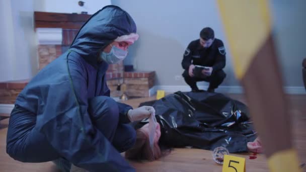 Portrait of police investigator putting spoon into transparent package. Officer using tablet at the background. Covered murder victim lying on the floor. Detectives gathering evidence at crime scene. — Stock Video