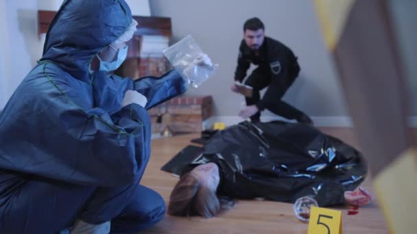 Kriminologe in Uniform zeigt einem Polizisten im Hintergrund einen Löffel in durchsichtiger Verpackung. Kriminalbeamter und Kriminaltechniker besprechen Spuren am Tatort. — Stockvideo