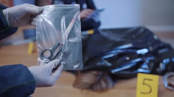 Close-up of blooded scissors in transparent package. Forensic investigator holding evidence at the crime scene. Covered murdered body lying on the floor at the background. — Stock Video