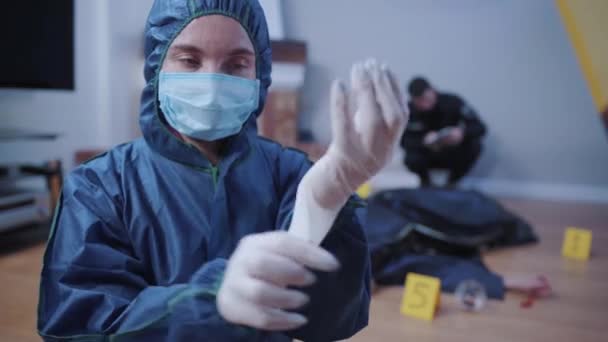 Retrato del investigador forense en uniforme poniéndose guantes blancos y mirando a la cámara. Una profesional que trabaja en la escena del crimen. Oficial de policía sentado al lado de la víctima en el fondo . — Vídeo de stock