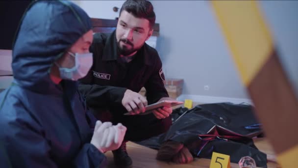 Porträtt av skäggiga unga kaukasiska polis som håller tablett och prata med kriminaltekniska utredare. Professionella som diskuterar brott bredvid mördade kroppar. Utredning, brottsbekämpning. — Stockvideo