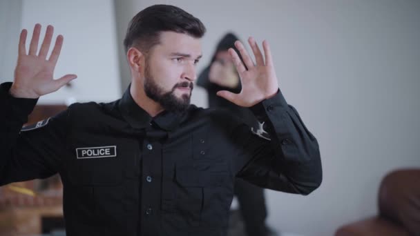 Scared Caucasian police officer standing with hands raised at the foreground, blurred criminal pointing gun on him from the background. Detective caught by robber at the crime scene. — Stock Video