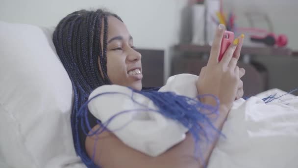 Портрет афроамериканской девушки с дредами, лежащими в белой кровати и использующей смартфон. Молодая женщина занимается серфингом по ночам. Зависимость от социальных сетей, стиль жизни . — стоковое видео