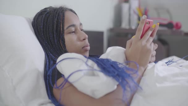 Chica afroamericana con rastas usando un teléfono inteligente, guardarlo y mirar hacia arriba pensativamente. Adolescente usando las redes sociales a la hora de acostarse. Adicción a Internet, estilo de vida . — Vídeos de Stock