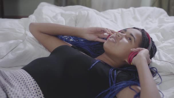十几岁的非洲裔美国女孩，带着可怕的头发，在耳机里听音乐，拿着智能手机，换着歌。 积极的女人躺在白色的床上。 休闲、休息、生活方式. — 图库视频影像