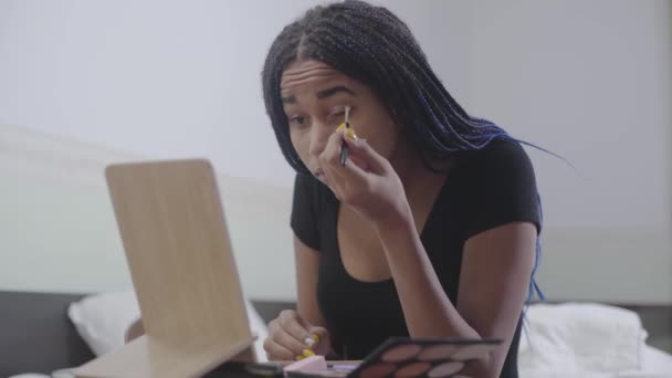 Porträtt av söt afroamerikansk tonårstjej som applicerar ögonskuggor. Ung kvinna med dreadlocks tittar på spegeln och gör upp sitt ansikte. Skönhet, ansiktsvård, livsstil. — Stockvideo