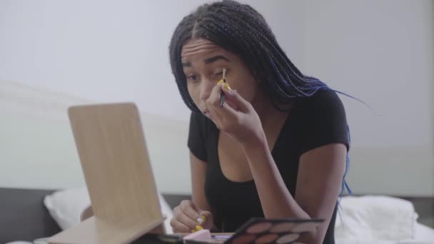 Retrato de uma adolescente afro-americana a aplicar sombras. Uma jovem com dreadlocks a olhar para o espelho e a inventar o rosto. Beleza, cuidado do rosto, estilo de vida . — Vídeo de Stock