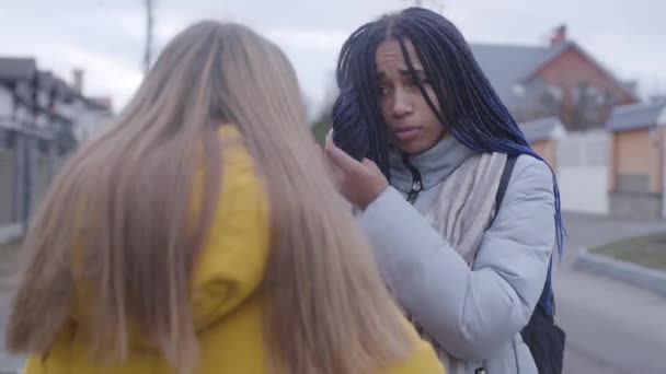 Zwei Freundinnen unterhalten sich, wenden sich einander zu, afrikanisch-amerikanischer Teenager geht, kaukasisches Mädchen steht mit ironischem Gesichtsausdruck. Streit, Lebensstil, Freundschaft. — Stockvideo