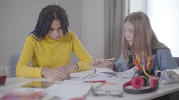 İçeride birlikte çalışan iki arkadaş. Afro-Amerikalı ve beyaz kızlar masada oturup yazı yazıyor. Eğitim, zeka, yaşam tarzı. — Stok video
