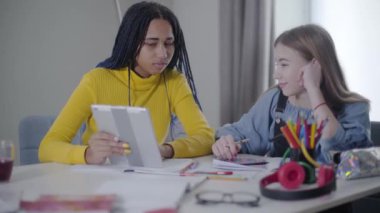 Rastalı Afro-Amerikalı kız beyaz arkadaşına tablet ekranda bir şey gösteriyor. Mutlu gençler evde ev ödevi yaparken dinleniyorlar. Zeka, öğrenim, eğitim, yaşam tarzı.