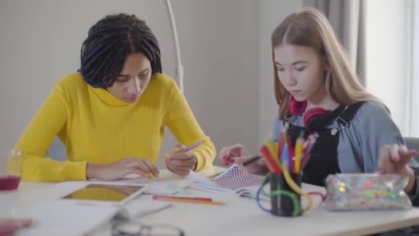 Retrato de dos alumnas haciendo deberes. Chicas caucásicas y afroamericanas sentadas a la mesa y escribiendo. Inteligencia, aprendizaje, estudio, educación . — Vídeo de stock