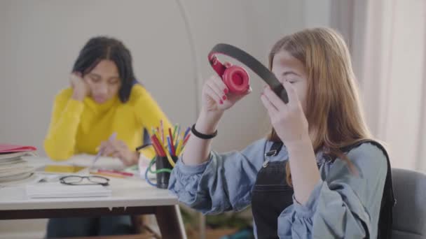 Chica bastante caucásica poniéndose los auriculares y escuchando música como su amigo afroamericano leyendo en el fondo. Los estudiantes pasan tiempo en el interior. Educación, ocio, estilo de vida . — Vídeos de Stock