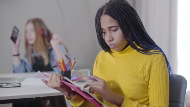 Estudante afro-americana cansada estudando como sua amiga caucasiana dançando em fones de ouvido ao fundo. Menina exausta segurando a cabeça com as mãos. Educação, problemas de comunicação . — Vídeo de Stock