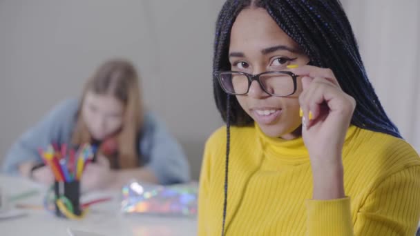 Close-up of beautiful African American woman looking at camera and holding eyeglasses. Cute girl in yellow sweater posing while her Caucasian friend studying at the background. — Stock Video